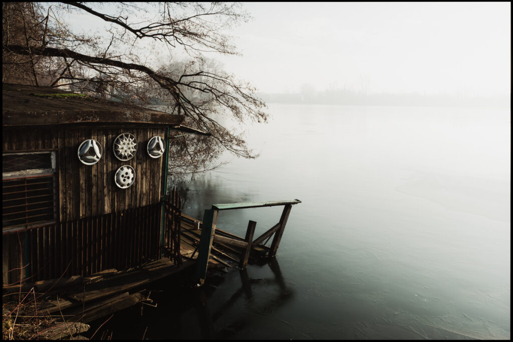 A rustic, wooden fishing pier and hut on the edge of the frozen, foggy Kopalniok pond near the Silesia mine, its wall strangely decorated with four silver car hubcaps.