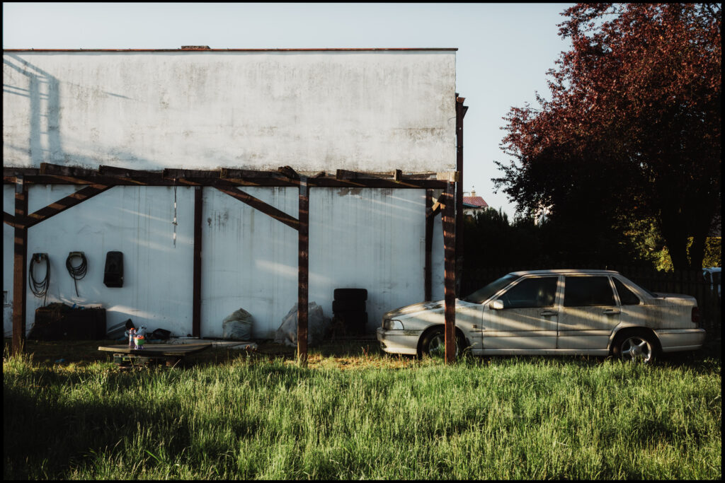 An old, dusty sedan parked on a grassy yard next to a stark white building, illuminated by the long shadows of a late afternoon sun.