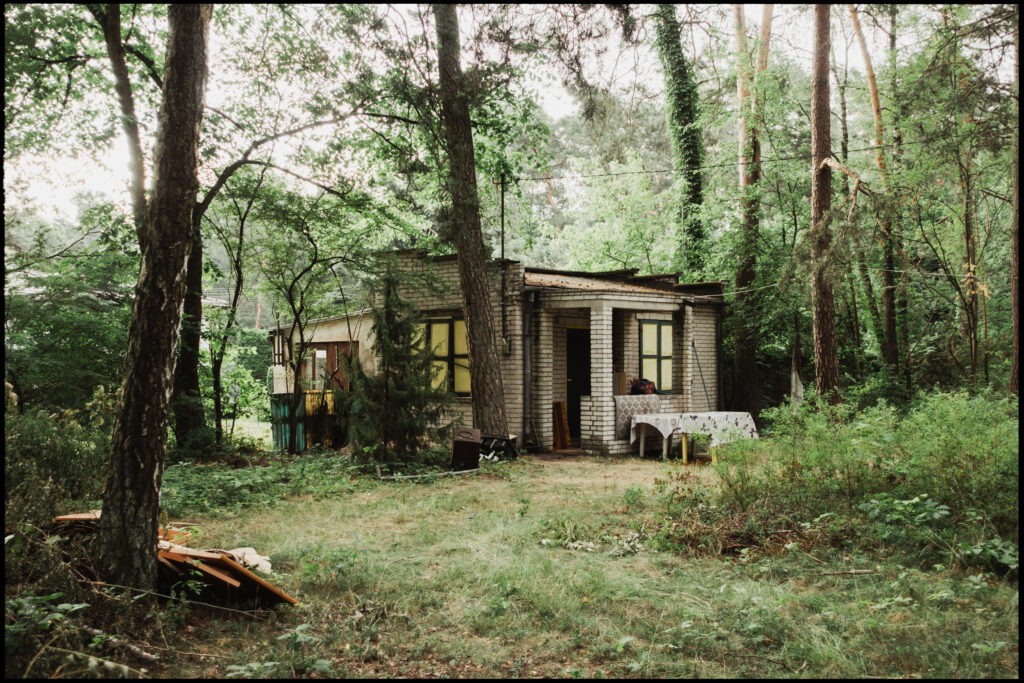 A small, brick-and-wood summer cottage in Urle, with yellow shutters and a makeshift porch, surrounded by an overgrown clearing in the woods.