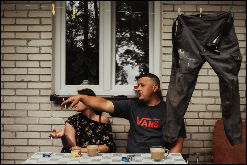 A candid photo of a man and a woman sitting at a table outside, drinking coffee and smoking cigarettes. The man is gesturing with his hand, while a pair of dirty work pants hangs on a clothesline next to him.