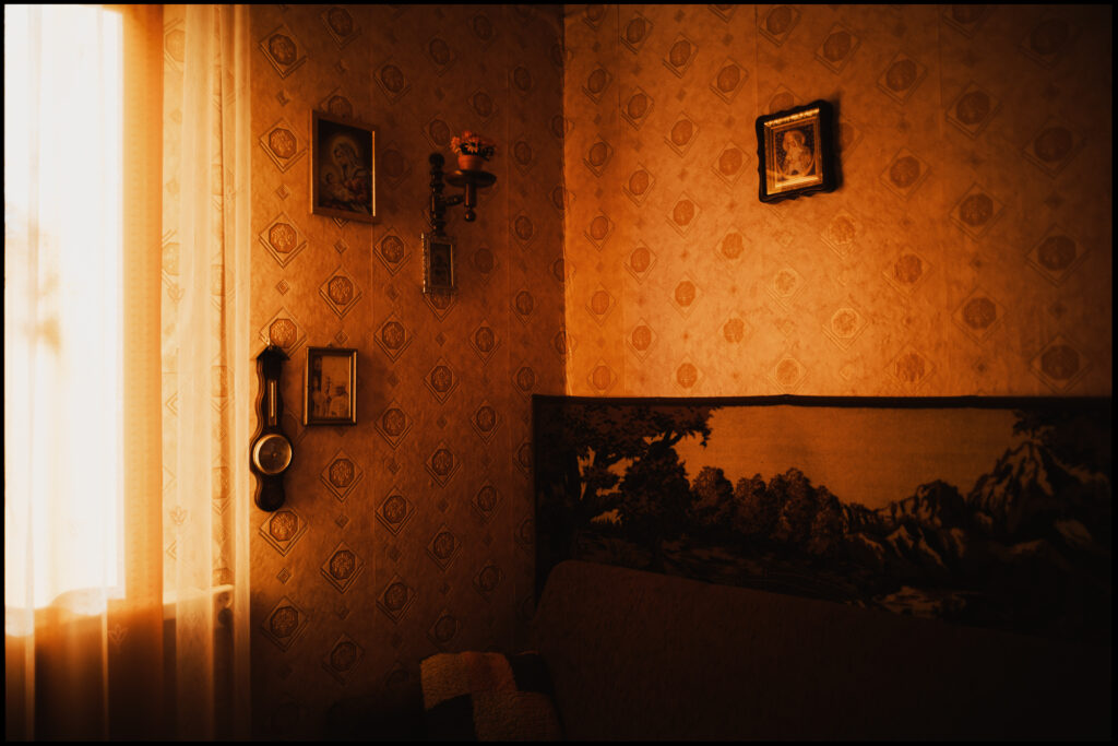 The interior of an abandoned house in Urle, a time capsule of a Polish home from a past era, with warm light filtering through a lace curtain onto vintage wallpaper and framed pictures.