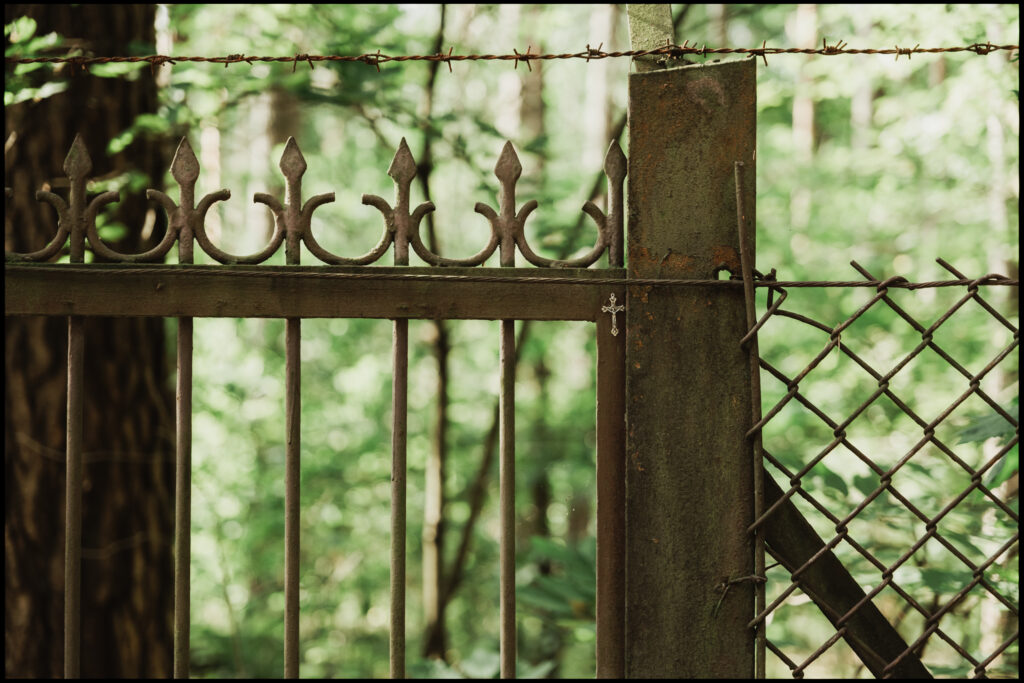 A close-up of a rusty, ornate metal gate topped with barbed wire, with a small, delicate cross hanging from it – an entrance to an abandoned property.