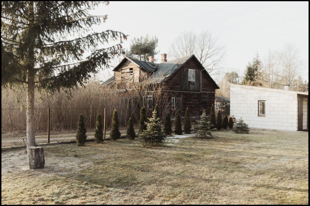 A large, dilapidated two-story wooden house with a damaged roof, standing next to a stark, modern white cinder block building in a frosty winter yard.