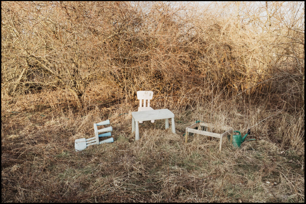 Abandoned children's furniture – a small white table and chair – left in a field of dry, overgrown winter grass and tangled bushes.