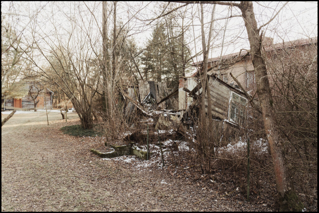 The collapsed ruins of a wooden structure, with broken walls and beams sticking out from a pile of rubble, surrounded by bare trees in winter.