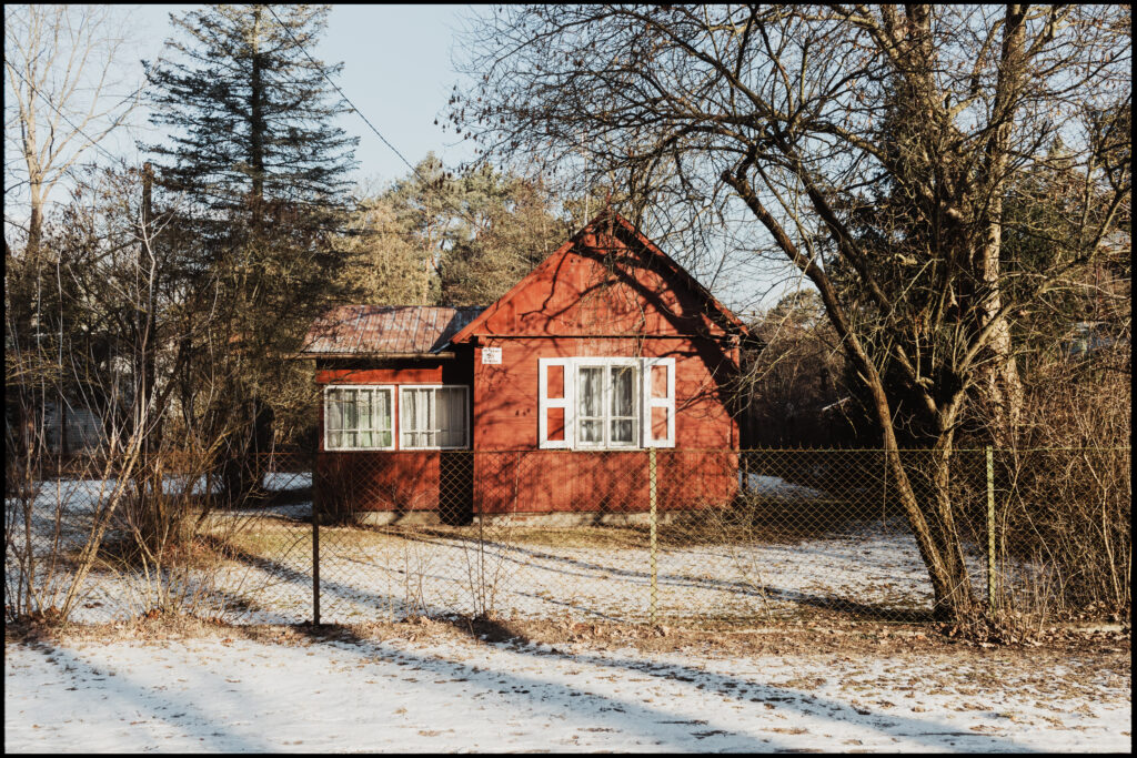 A small, bright red wooden cottage with white window frames, seen behind a wire fence on a snowy winter day.