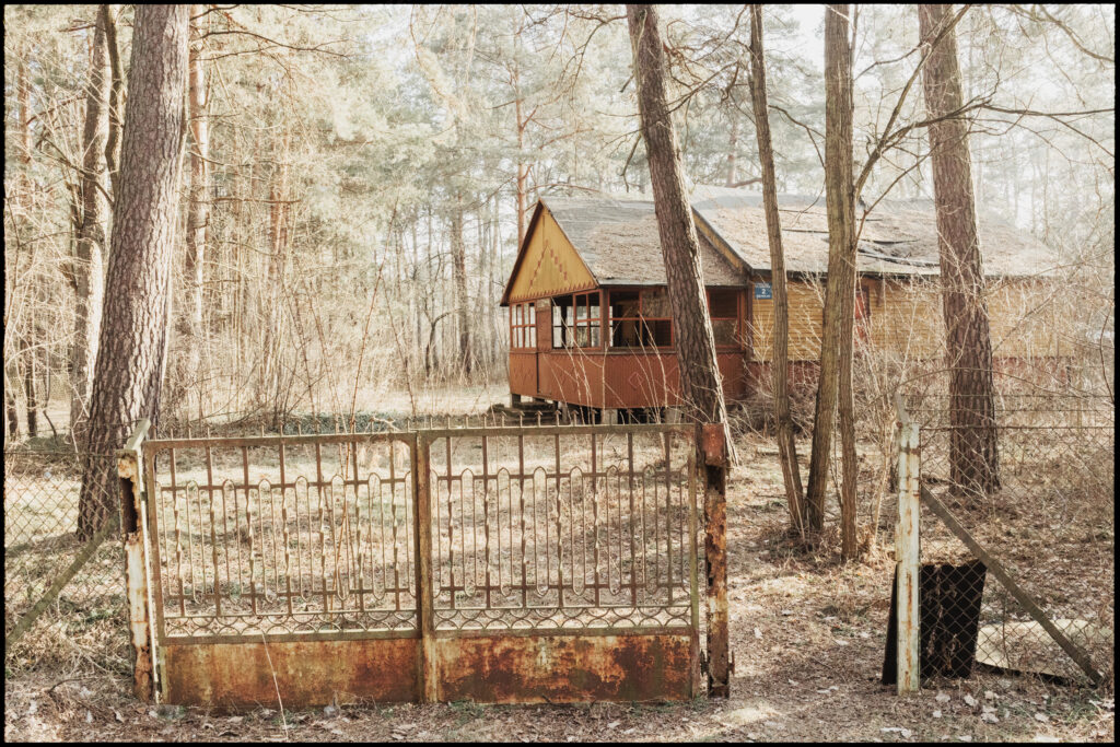 A rusty, wrought-iron gate stands ajar in front of a weathered, yellow wooden cottage in Urle during winter, with bare trees and a washed-out, hazy light.