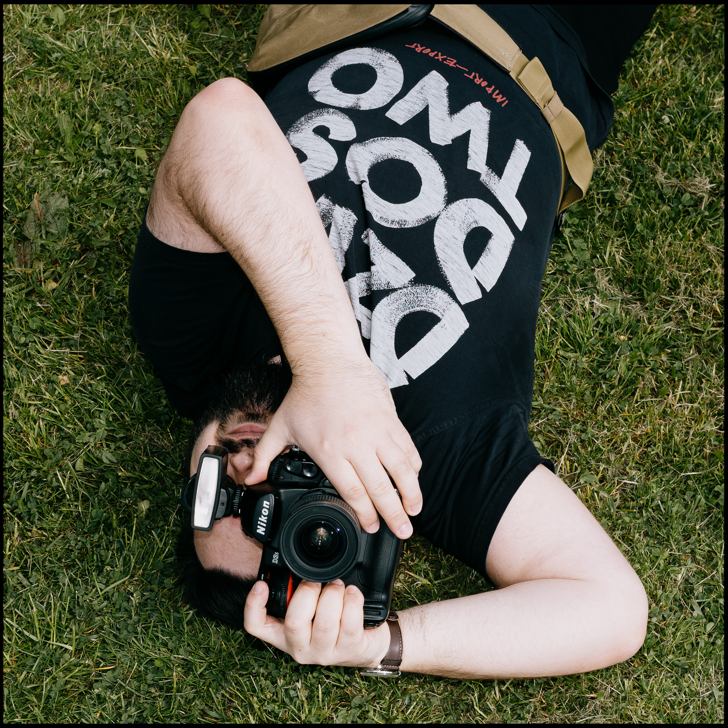 A top-down portrait of Paweł Świątek, the photographer from the SaintyLtd 'about me' page, lying on the grass and holding his Nikon camera.