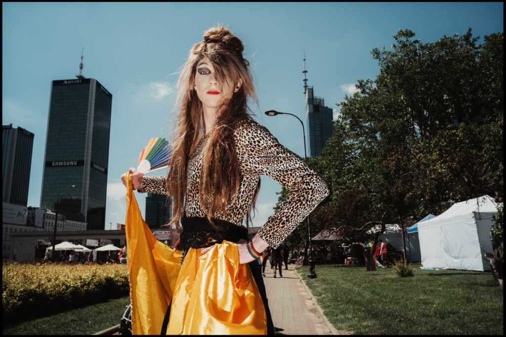 A drag queen strikes a confident pose on a sidewalk at Warsaw Pride 2024, wearing a leopard print top, a flowing yellow skirt, and holding a rainbow fan.