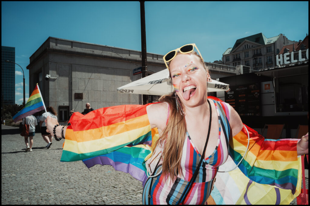A woman with a fun, defiant expression sticks her tongue out at the camera, wearing a large rainbow flag as a cape and colorful glitter makeup during Warsaw Pride 2024.