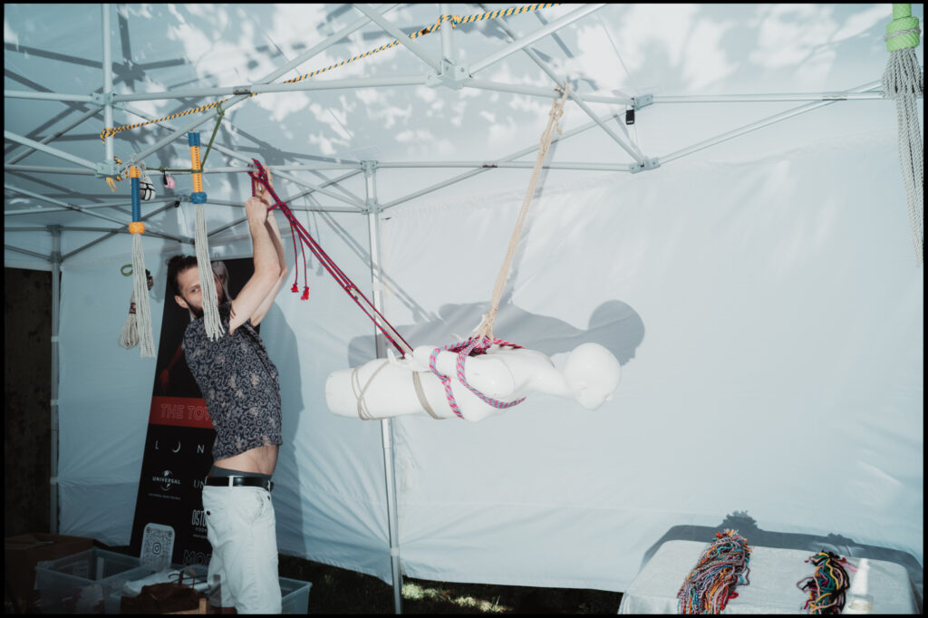A man demonstrates Japanese shibari rope art, adjusting the suspension ropes on a white mannequin inside a tent at a Warsaw Pride 2024 booth.