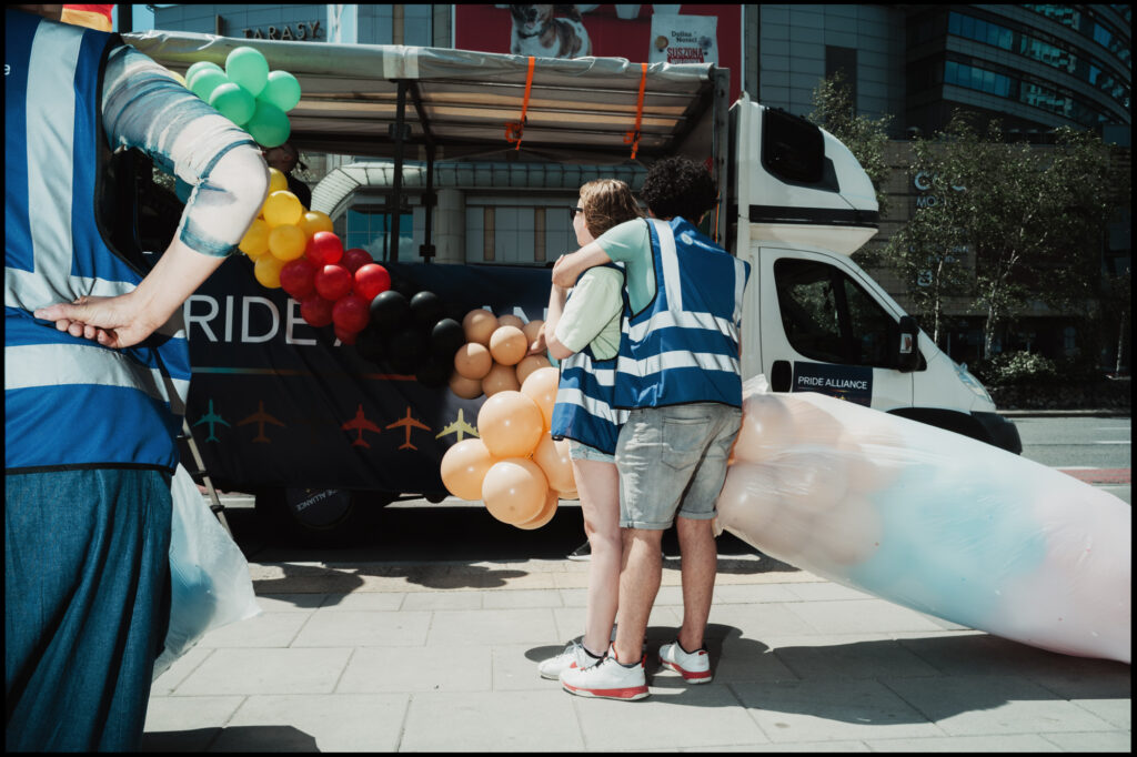 Two volunteers in safety vests share a warm hug next to a parade truck decorated with rainbow and trans-colored balloons for Warsaw Pride 2024.