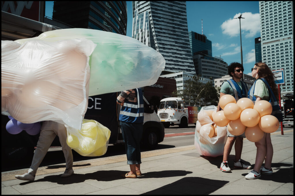 Participants carry large, pastel-colored inflatables and bunches of orange balloons down a city street during the Warsaw Pride 2024 parade.