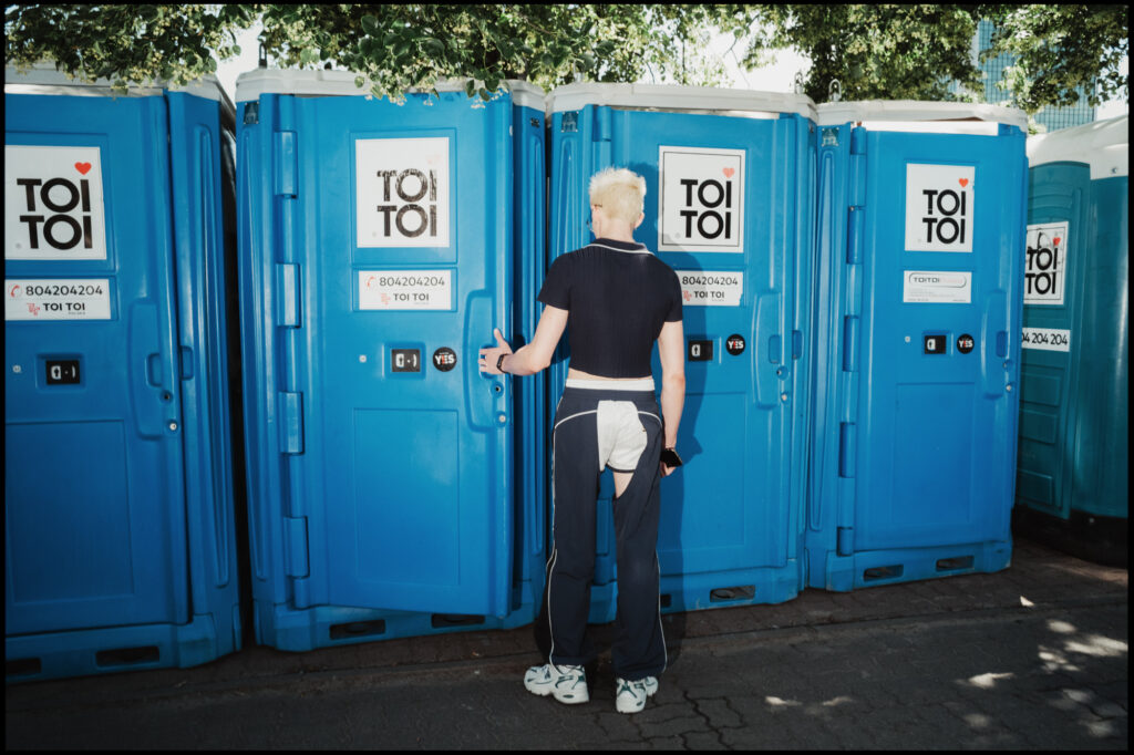A candid, slice-of-life moment from Warsaw Pride 2024: a participant with blonde hair stands in front of a row of blue TOI TOI portable toilets.