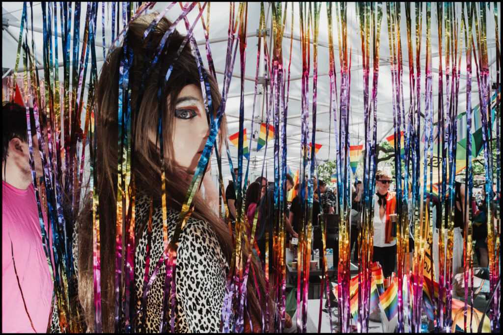 A drag queen in a leopard print top with dramatic eye makeup peeks through a curtain of colorful metallic streamers at the Warsaw Pride event.
