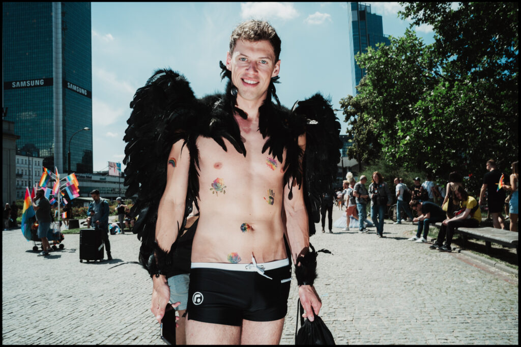 A smiling man wearing large black feathered wings and small rainbow tattoos on his chest poses directly for the camera in a sunny city square during Warsaw Pride 2024.