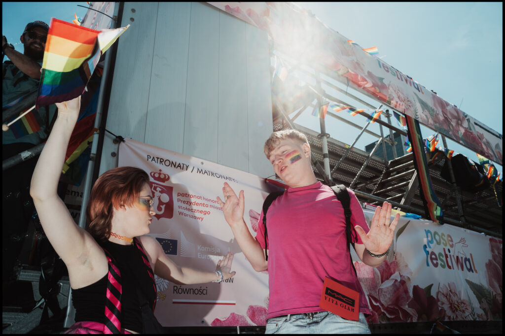 A bright, sun-flared photo of two participants dancing and celebrating by a parade truck during Warsaw Pride 2024, one holding a rainbow flag high.