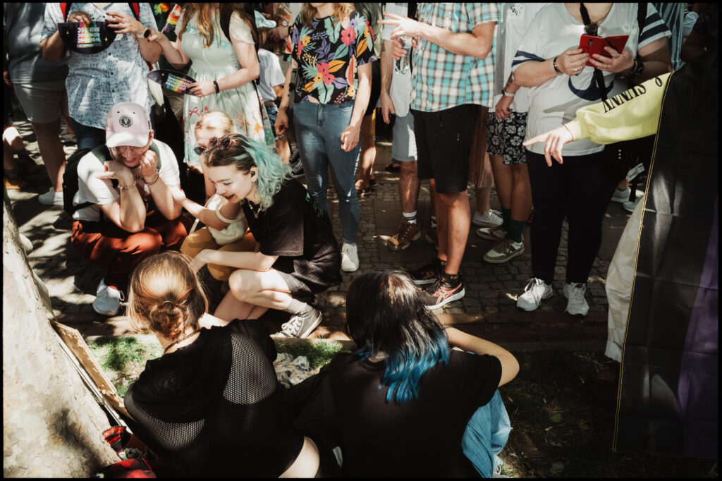 A candid moment of community at Warsaw Pride 2024, where a group of young people are resting in the shade. In the center, two attendees crouch down, sharing a joyful, smiling interaction.