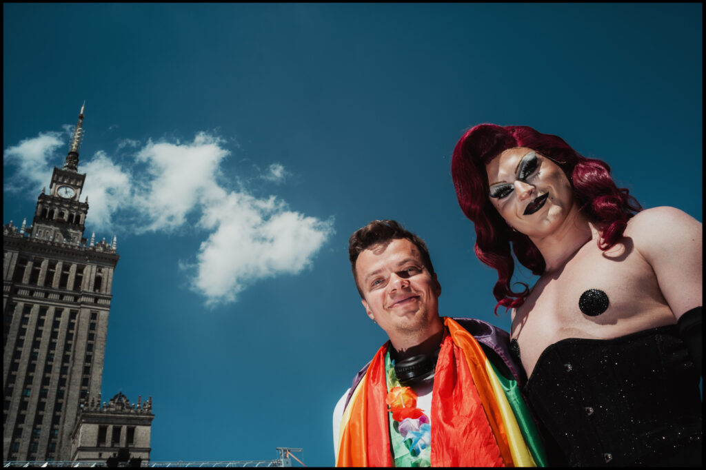 A dramatic low-angle shot of a smiling man and a drag queen with a red wig, posing against a bright blue sky with Warsaw's Palace of Culture behind them during Warsaw Pride 2024.