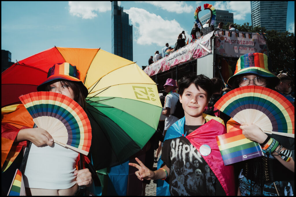 Three young participants at Warsaw Pride 2024 pose for the camera, holding colorful rainbow fans and a rainbow umbrella, with one making a peace sign.