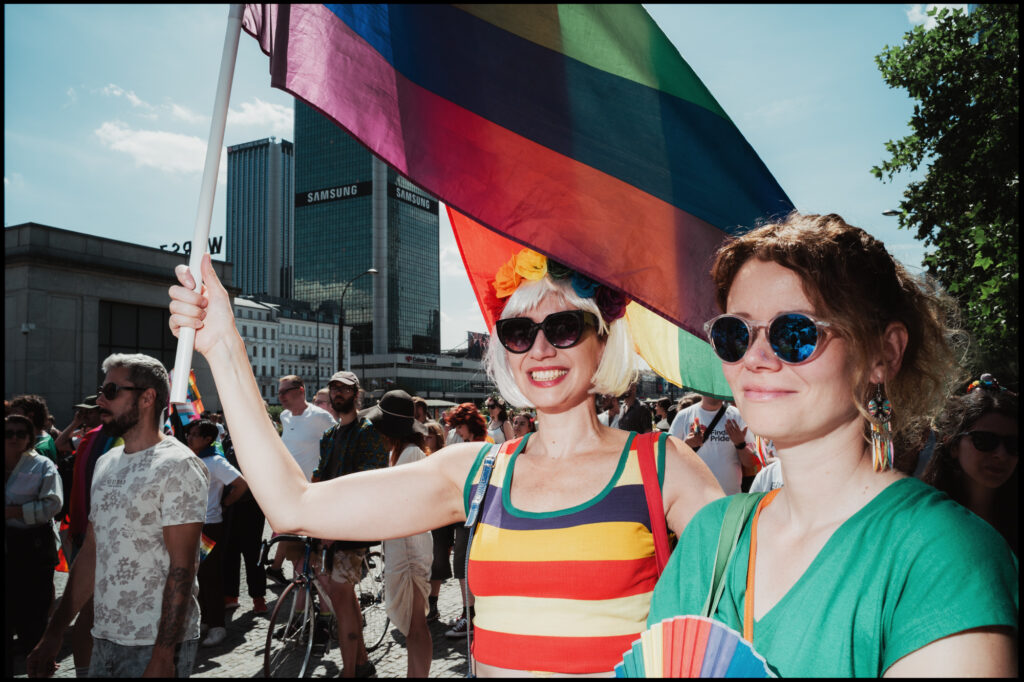 Two smiling women in sunglasses stand in the crowd at Warsaw Pride 2024, one proudly hoisting a large rainbow flag against the city skyline.