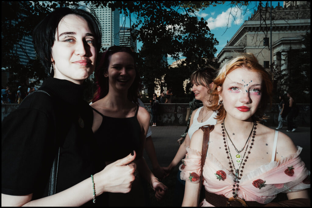 A group of young friends at Warsaw Pride 2024; one woman in the foreground has creative makeup with facial gems and a pink strawberry-themed top.