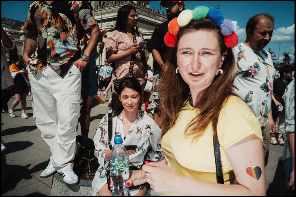 A candid portrait of a woman in a crowd at Warsaw Pride 2024, wearing a yellow shirt, a rainbow pom-pom headband, and a rainbow heart sticker on her arm.