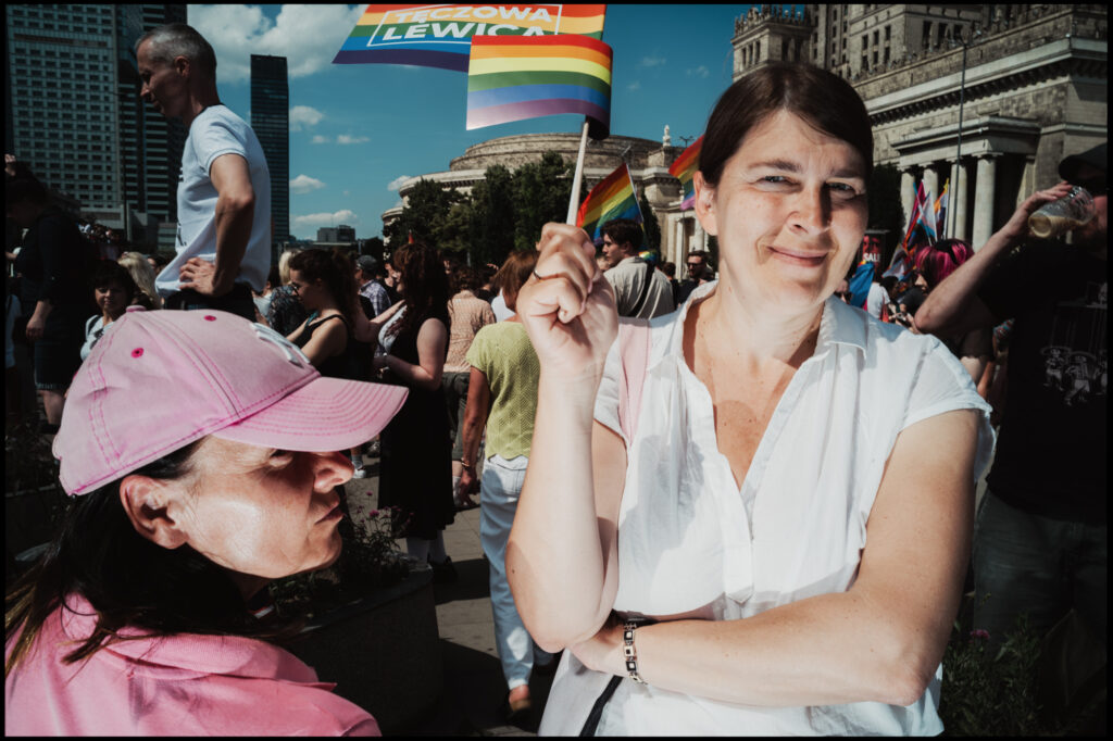 A woman with a gentle smile holds up a small rainbow flag, looking at the camera during the Warsaw Pride 2024 march, with another attendee in a pink hat in the foreground.