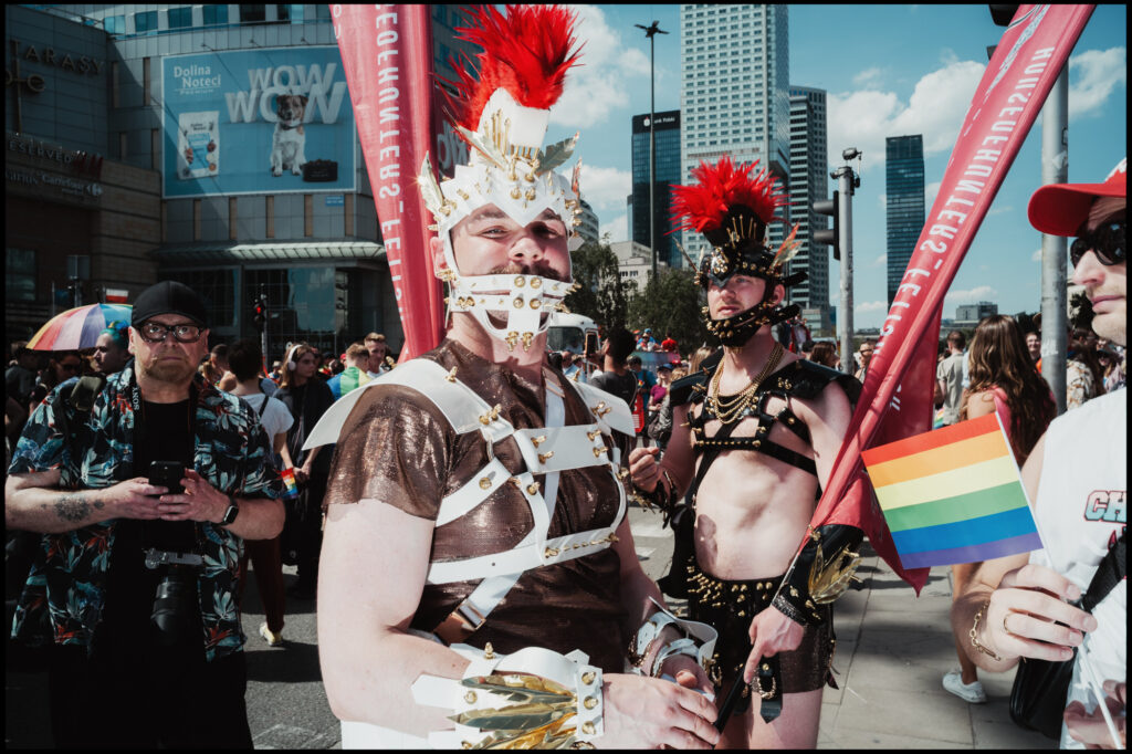 A participant in an elaborate Roman gladiator-style costume, complete with a red-plumed helmet and leather harness, marches in the crowd at Warsaw Pride 2024 holding a rainbow flag.