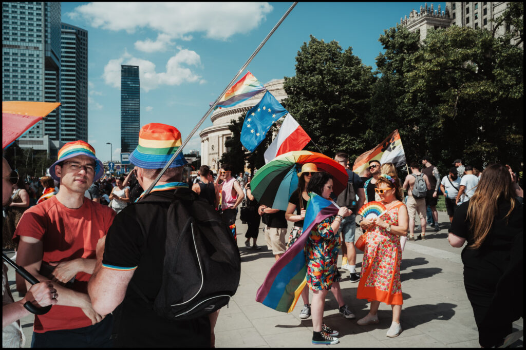 A wide, sunny street scene from Warsaw Pride 2024, showing a diverse crowd of people marching, carrying various rainbow flags, and enjoying the parade.