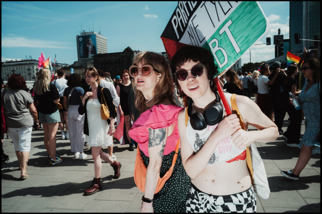 Two friends march at Warsaw Pride 2024; the person in front, wearing checkered pants and sunglasses, holds up a green protest sign and smiles at the camera.