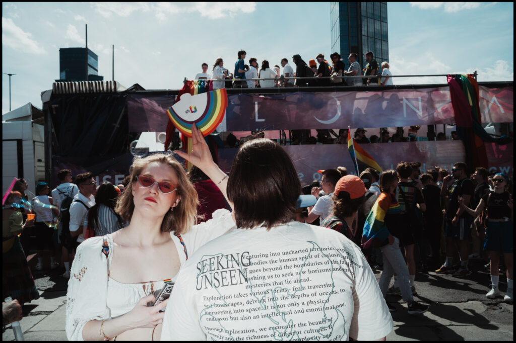 A stylish couple watches a parade float pass by at Warsaw Pride 2024, with the sun creating a bright flare in the background.