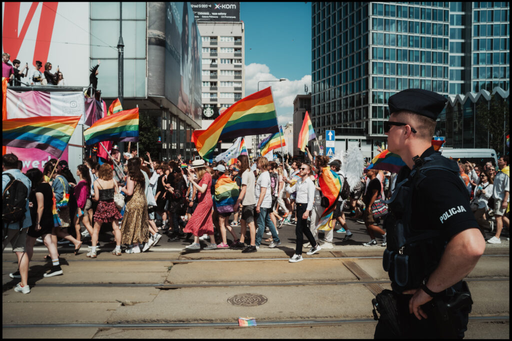 A Polish police officer stands in the foreground, observing the colorful and energetic crowd marching with rainbow flags down a city street during Warsaw Pride 2024.