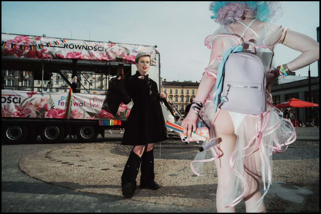 Two attendees at Warsaw Pride 2024 show off contrasting styles: one in a dark, gothic outfit and the other in a light, pastel-colored costume with a lavender backpack.