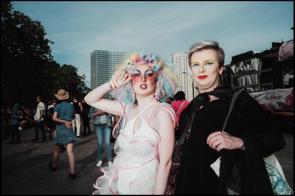 A street style portrait of two participants at Warsaw Pride 2024: one wears an elaborate pastel rainbow wig and white outfit, while the other poses confidently with silver hair and bold makeup.