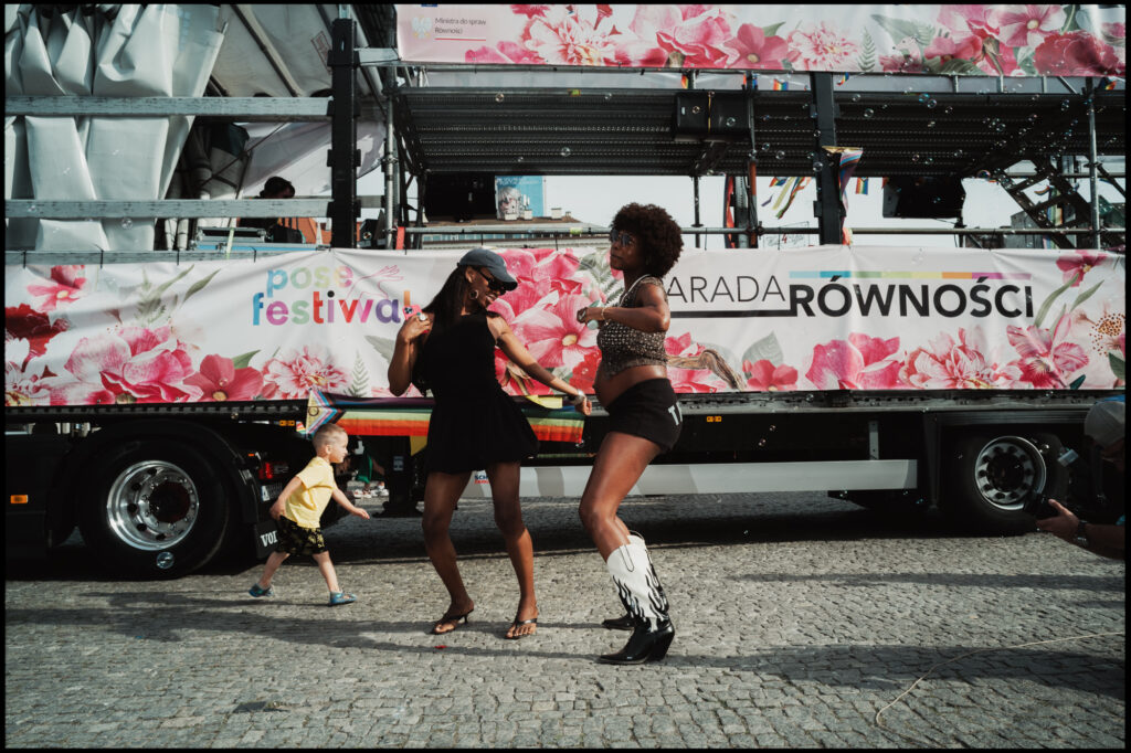 Two women joyfully dance on a cobblestone street in front of a parade truck with a "Parada Równości" banner, as a small child in a yellow shirt runs past in the background during Warsaw Pride 2024.