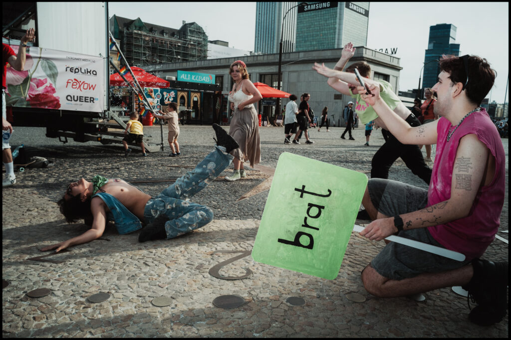 A theatrical scene at Warsaw Pride 2024: one person lies on the pavement while another kneels, pointing and holding a green sign with the word "brat".