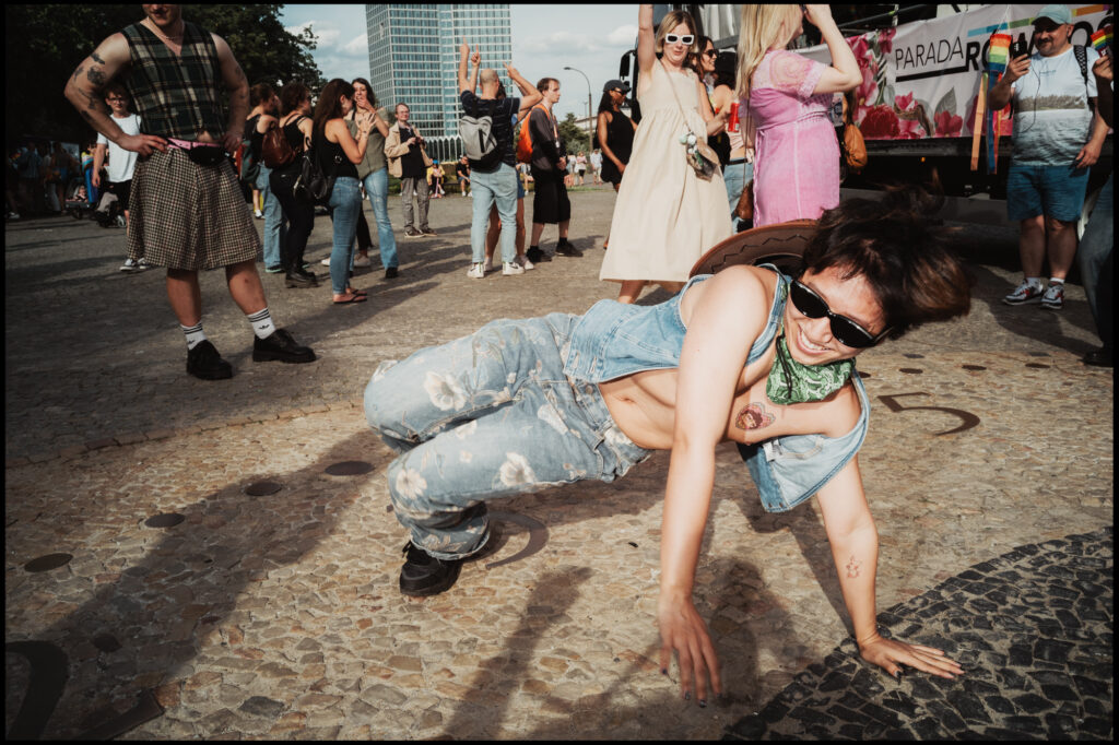 A participant in a denim outfit performs a dynamic dance move, flipping their long dark hair in the bright sun during Warsaw Pride 2024.