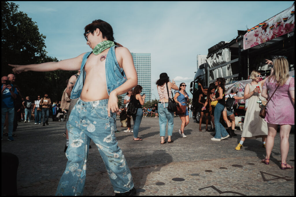 A joyful dancer in a denim vest and printed jeans points to the side while moving to the music on a cobblestone street at Warsaw Pride 2024.