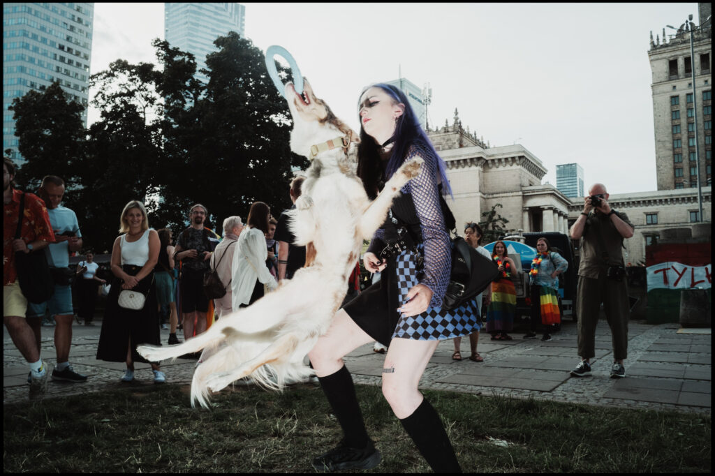A dynamic scene from Warsaw Pride 2024: a participant with purple hair and a checkered skirt plays with her dog, which is captured gracefully leaping through the air in a park.